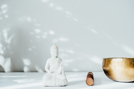 Decorative white Buddha statuette and tibetian singing bowl on the white background with sun light shadows. Meditation and relaxation ritual. Exotic massage. Minimalism. Copy space. Selective focus.